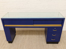 Load image into Gallery viewer, A mid-century modern blue desk with a glass top, gold metal accents, and three drawers on the right side, set against a white brick wall and beige carpet.
