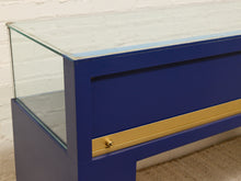 Load image into Gallery viewer, A close-up of a modern display case or console table with a deep blue painted frame, a clear glass top, and a horizontal brass handle.
