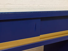 Load image into Gallery viewer, Close-up of a blue wooden furniture piece with a gold metal rail and a small pull handle, set against a white brick wall.
