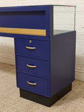 Load image into Gallery viewer, A vintage-style blue display cabinet with three drawers, gold handles, a black base, and a glass top, set against a white brick wall and beige carpet.
