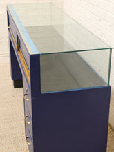 Load image into Gallery viewer, A navy blue wooden display case or console table with a clear glass top, gold drawer pulls, and a light blue protective film along the glass edge, set against a white brick wall and beige carpet.
