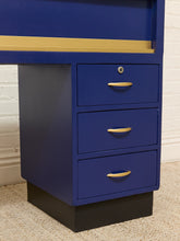 Load image into Gallery viewer, A close-up of a blue three-drawer pedestal cabinet with gold handles and a black base, shown against a white brick wall and beige carpet.
