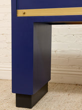 Load image into Gallery viewer, Close-up of a modern desk leg featuring a deep blue painted finish, a gold horizontal accent strip, and a black block base, set against a white brick wall and beige carpet.
