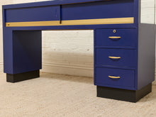 Load image into Gallery viewer, A mid-century modern writing desk in royal blue with gold metal drawer pulls and a gold horizontal accent strip, set against a white brick wall and beige carpet.
