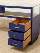 Load image into Gallery viewer, A blue and wood display cabinet with three open drawers, a glass top shelf, and gold-tone handles, set against a white brick wall and beige carpet.
