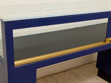 Load image into Gallery viewer, A close-up of a blue display case or cabinet with a glass front, featuring a gold metal handle and trim, set against a white brick wall and beige carpet.
