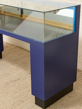 Load image into Gallery viewer, A modern jewelry display case with a deep blue painted metal frame, clear glass top and sides, and a black base, shown on a beige carpeted floor.
