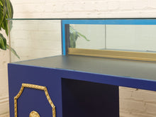 Load image into Gallery viewer, A blue display case or console table with ornate gold trim, a dark gray top surface, and a clear glass front panel, shown against a white brick wall.
