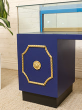 Load image into Gallery viewer, A modern blue display case or cabinet with ornate gold trim and a central gold medallion, set on a black base in a room with a white brick wall and beige carpet.
