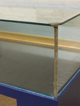 Load image into Gallery viewer, Close-up of a glass display case corner with a clear glass top and front panel, a gold-toned inner trim, and a dark blue base.
