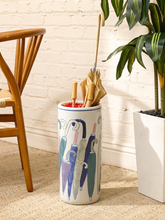 Load image into Gallery viewer, A cylindrical ceramic umbrella stand with a white base and a colorful painted umbrella pattern, holding several umbrellas. It is styled in a home entryway setting next to a wooden chair and a potted plant against a white brick wall.
