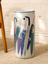 Load image into Gallery viewer, A tall cylindrical ceramic umbrella stand with a white glaze, decorated with a pattern of colorful hand-drawn umbrellas in shades of green, blue, purple, and beige. The stand is photographed on a textured beige rug in front of a white brick wall, with a wooden chair leg visible on the left.
