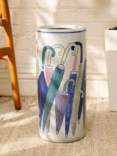 Load image into Gallery viewer, A tall cylindrical ceramic umbrella stand with a white glaze, decorated with a colorful illustration of multiple umbrellas in shades of blue, green, purple, and beige. The stand is photographed on a textured beige rug in front of a white brick wall, with a wooden chair leg visible on the left.
