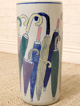 Load image into Gallery viewer, A tall cylindrical ceramic vase or planter with a white glaze, decorated with a colorful illustration of overlapping umbrellas in blue, green, yellow, and purple. The vase is photographed standing on a beige woven rug against a white brick wall background.
