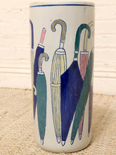 Load image into Gallery viewer, A tall cylindrical ceramic container or vase with a hand-painted pattern of colorful umbrellas on a white background, set against a light brick wall and textured beige surface.
