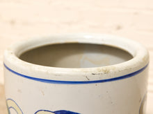 Load image into Gallery viewer, Close-up of a white ceramic stoneware container or crock with a blue painted rim stripe and partial blue decorative motif, set against a light wood background.
