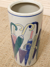 Load image into Gallery viewer, A white ceramic cylindrical container or vase decorated with a colorful hand-painted umbrella pattern, resting on a beige woven rug.

