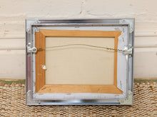 Load image into Gallery viewer, The back view of a square framed canvas print, showing a silver metal frame, wooden inner border, backing board, and hanging wire against a cream wall and woven rug.
