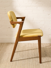 Load image into Gallery viewer, Image 74 of 89: A mid-century modern wooden dining chair with a light yellow upholstered seat and backrest, positioned against a white brick wall on a textured beige rug.
