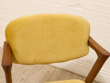 Load image into Gallery viewer, Image 77 of 89: A close-up of a mid-century modern armchair featuring a mustard yellow velvet upholstered backrest and wooden arms, set against a white brick wall and light-colored rug.

