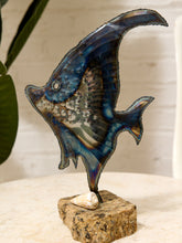 Load image into Gallery viewer, A stained glass sculpture of an angelfish mounted on a stone base with a small shell, displayed on a tabletop against a light wall with a plant leaf in the background.
