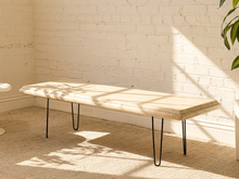 Load image into Gallery viewer, A modern bench with a light beige stone top and black hairpin legs, positioned in a room with a white brick wall and beige carpet, lit by warm natural sunlight casting leaf shadows.
