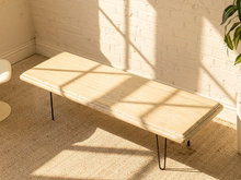 Load image into Gallery viewer, A modern rectangular bench with a light beige travertine stone top and black metal hairpin legs, styled in a room with a white brick wall and warm sunlight casting windowpane shadows.
