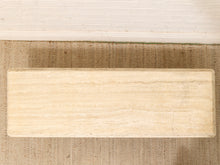 Load image into Gallery viewer, A rectangular beige stone tile or slab with natural horizontal veining, shown from a top-down view on a woven brown rug against a white brick wall.
