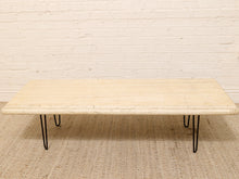 Load image into Gallery viewer, A rectangular coffee table with a light beige travertine stone top and black hairpin legs, set on a woven jute rug against a white brick wall.
