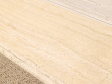 Load image into Gallery viewer, A close-up of a light beige stone surface with natural veining, showing part of a matching stone edge and a white brick wall background.
