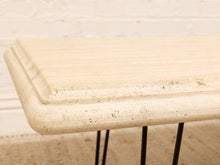 Load image into Gallery viewer, Close-up of a cream-colored travertine stone tabletop with layered beveled edges, supported by slim black metal hairpin legs against a light brick wall background.
