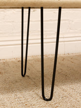 Load image into Gallery viewer, Close-up of black hairpin legs supporting a light beige stone or wood tabletop, standing on a textured beige rug against a white brick wall.
