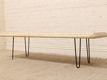 Load image into Gallery viewer, A modern bench with a light beige travertine stone top and black hairpin metal legs, positioned against a white brick wall and beige carpet.
