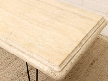Load image into Gallery viewer, Close-up of a cream-colored travertine stone tabletop with a textured natural stone surface, featuring a raised beveled edge and supported by thin black metal hairpin legs, set on a woven jute rug.
