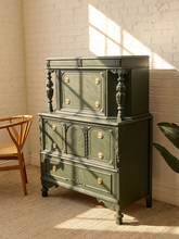 Load image into Gallery viewer, An antique-style three-tier chest of drawers painted in dark olive green, featuring brass knobs and turned legs, set against a white brick wall with warm sunlight and shadows.
