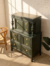 Load image into Gallery viewer, A dark green vintage-style hutch or cabinet with ornate gold-toned drawer knobs, shown in a sunlit room with a white brick wall, a wooden chair edge, and a plant leaf visible.
