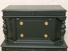 Load image into Gallery viewer, A dark forest green wooden chest of drawers with ornate gold drawer pulls, set against a white brick wall background.
