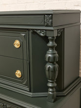 Load image into Gallery viewer, Close-up of a dark green carved wooden chest of drawers with ornate gold drawer pulls and a decorative turned leg, set against a white brick wall background.
