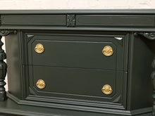Load image into Gallery viewer, A close-up of a dark green wooden cabinet with ornate gold drawer pulls, set against a white brick wall background.
