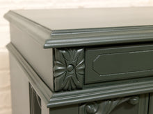 Load image into Gallery viewer, Close-up of a dark green painted wooden cabinet corner with decorative carved details and a drawer front, set against a light brick wall background.
