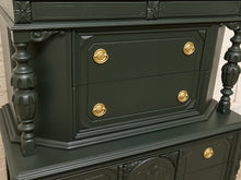 Load image into Gallery viewer, A close-up of a dark forest green wooden cabinet with ornate gold drawer knobs and carved details, set against a light brick wall background.
