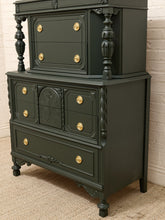 Load image into Gallery viewer, A dark forest green painted wooden chest of drawers with ornate gold drawer pulls, standing on a beige carpet against a white brick wall.
