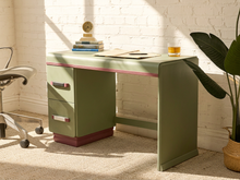 Load image into Gallery viewer, A retro-style writing desk in sage green with a dusty rose pink top edge and base, styled in a sunlit room with a white brick wall, office chair, and potted plant.
