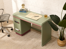 Load image into Gallery viewer, A sage green desk with pink trim and dark red base accents, styled in a home office setting with a white brick wall, a potted plant, and an office chair.
