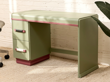 Load image into Gallery viewer, An olive green desk with a matching three-drawer filing cabinet, accented by a thin pink stripe along the top edge and a dark red base. The desk is set in a room with a white brick wall, beige carpet, and a leafy plant in the background.

