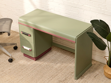 Load image into Gallery viewer, A retro-style writing desk in sage green with pink trim and drawer pulls, positioned in a home office setting next to an office chair and a potted plant.
