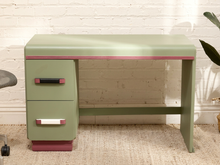 Load image into Gallery viewer, A retro-style writing desk in sage green with a matching pedestal drawer unit on the left, accented by a thin pink stripe along the top edge and drawer pulls with pink and white ends. The desk is positioned against a white brick wall on a beige rug.
