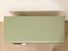 Load image into Gallery viewer, A minimalist rectangular coffee table with a solid sage green top, photographed from a top-down perspective on a woven jute rug against a white brick wall.
