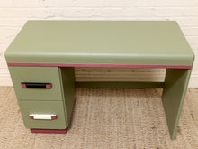 Load image into Gallery viewer, A mid-century modern writing desk with a sage green finish, featuring a pink stripe detail along the front edge, dual drawers on the left with black and white handles, and a pink base trim, set against a white brick wall and woven rug.
