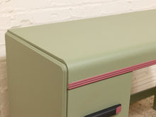 Load image into Gallery viewer, A close-up of a sage green desk with a pink decorative stripe detail and a black-and-pink drawer pull handle, set against a white brick wall background.
